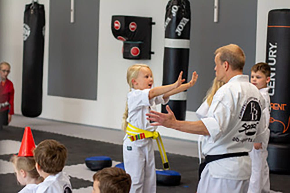 Kind beim Kampfsporttraining bei BB Sports: kleines Mädchen übt spielerisch eine Abwehrbewegung mit Trainer, im Hintergrund weitere Kinder und Trainingsgeräte.
